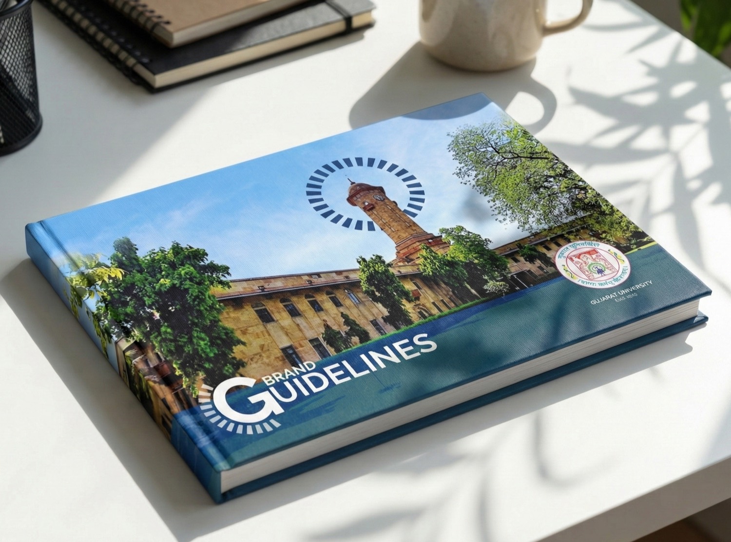 The Gujarat University brand guidelines book resting on a desk, clock-tower cover photograph and brand-device halo visible